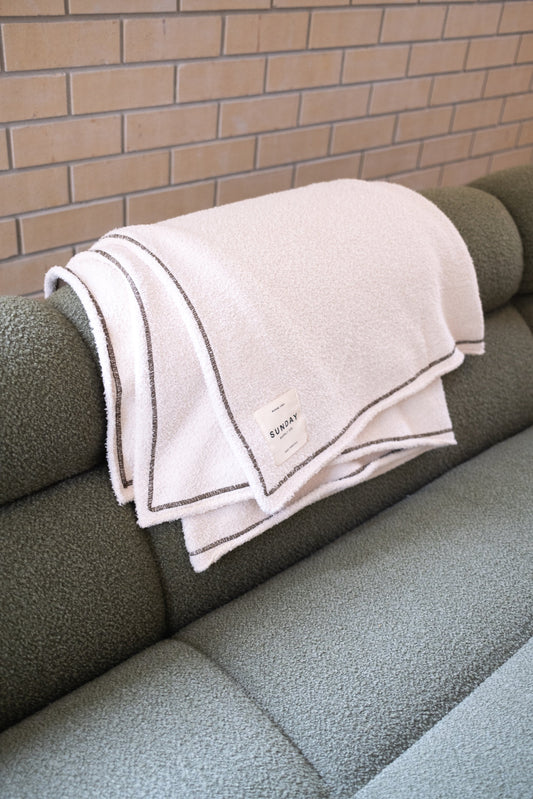 Cream Dunes knitted home blanket draped over the arm of a green sofa