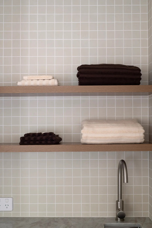 Folded Chocolate and Dunes towels styled on timber bathroom shelves against tile